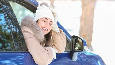 Frau sitzt mit Winterjacke, Haube und Handschuhe im Auto und lehnt sich aus dem Auto