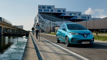 Blauer Renault ZOE fährt am Wasser entlang neben zwei Fahrrad-Fahrern